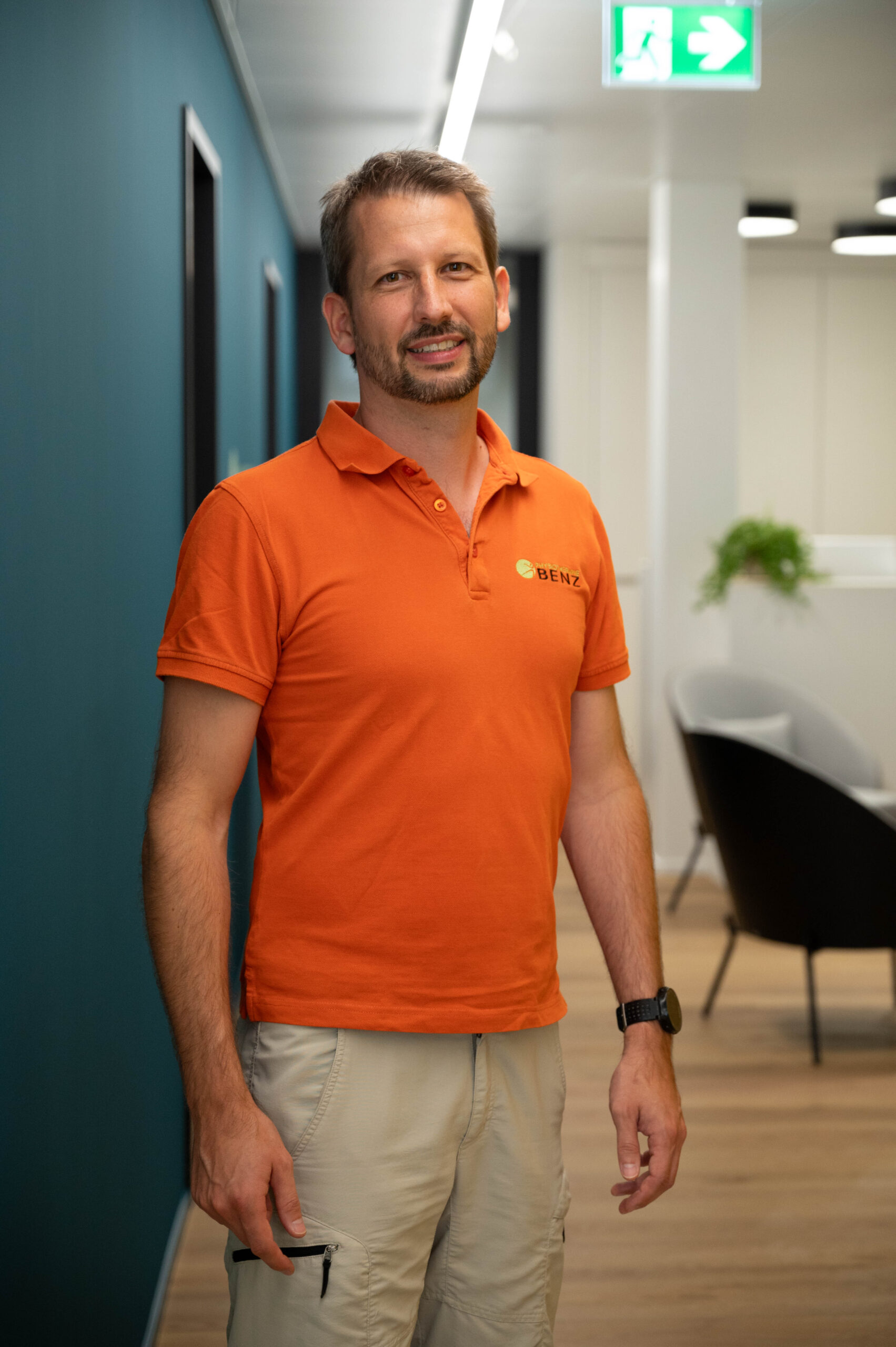 Simon Benz trägt ein oranges Poloshirt mit der Aufschrift „BENZ“ und beige Hosen. Er steht in einem Flur und lächelt in die Kamera. Hinter ihm sieht man ein paar Stühle und Pflanzen, und an der Decke hängt ein grünes Notausgangsschild.